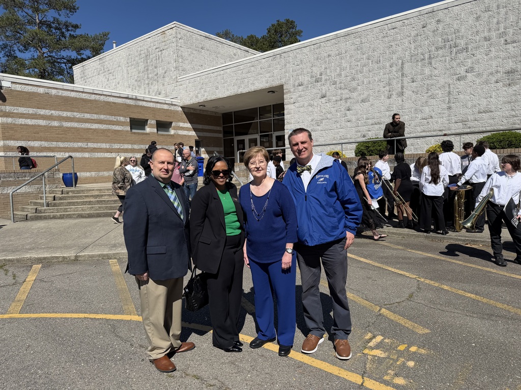 Four school leaders pose outside building as students and band members gather nearby under clear blue sky during event.