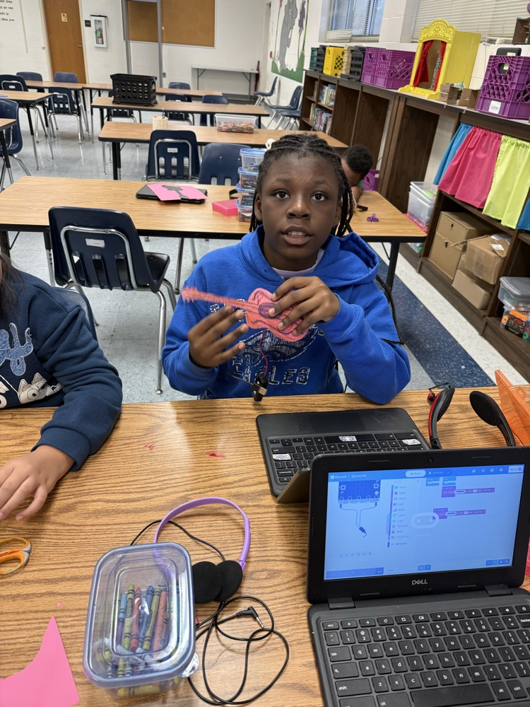 2. Two students sitting at a table smiling as one holds up a decorated paper guitar connected to a micro:bit, with laptops and crayons nearby