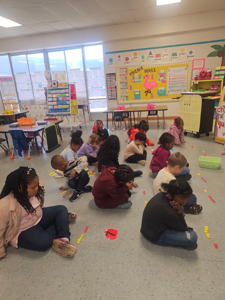 A group of kindergarten students sit on the classroom floor using colored counters to build different combinations of numbers during a hands-on math activity.