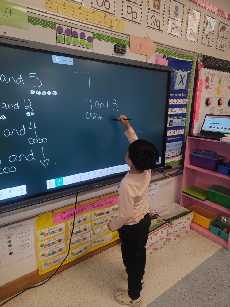 A young student stands at an interactive board writing number combinations for 7, using circles to represent quantities while solving a math problem.