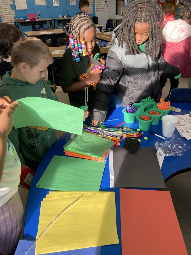 A group of elementary students gather around a table filled with green, yellow, and black construction paper, pipe cleaners, and craft supplies as they plan their leprechaun trap.