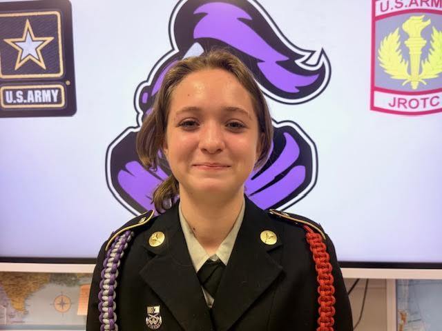 A smiling female JROTC cadet in a black dress uniform with purple and silver cords, posing in front of a U.S. Army JROTC backdrop