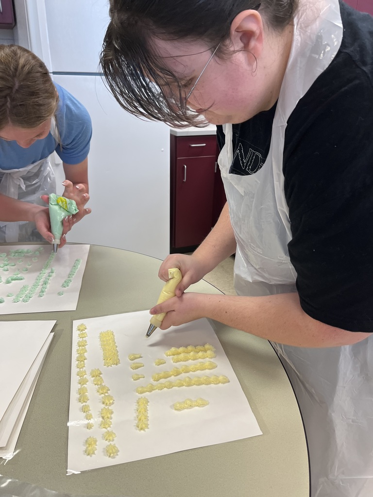 Two students in blue and black shirts wearing plastic aprons lean over a table, carefully decorating yellow treats