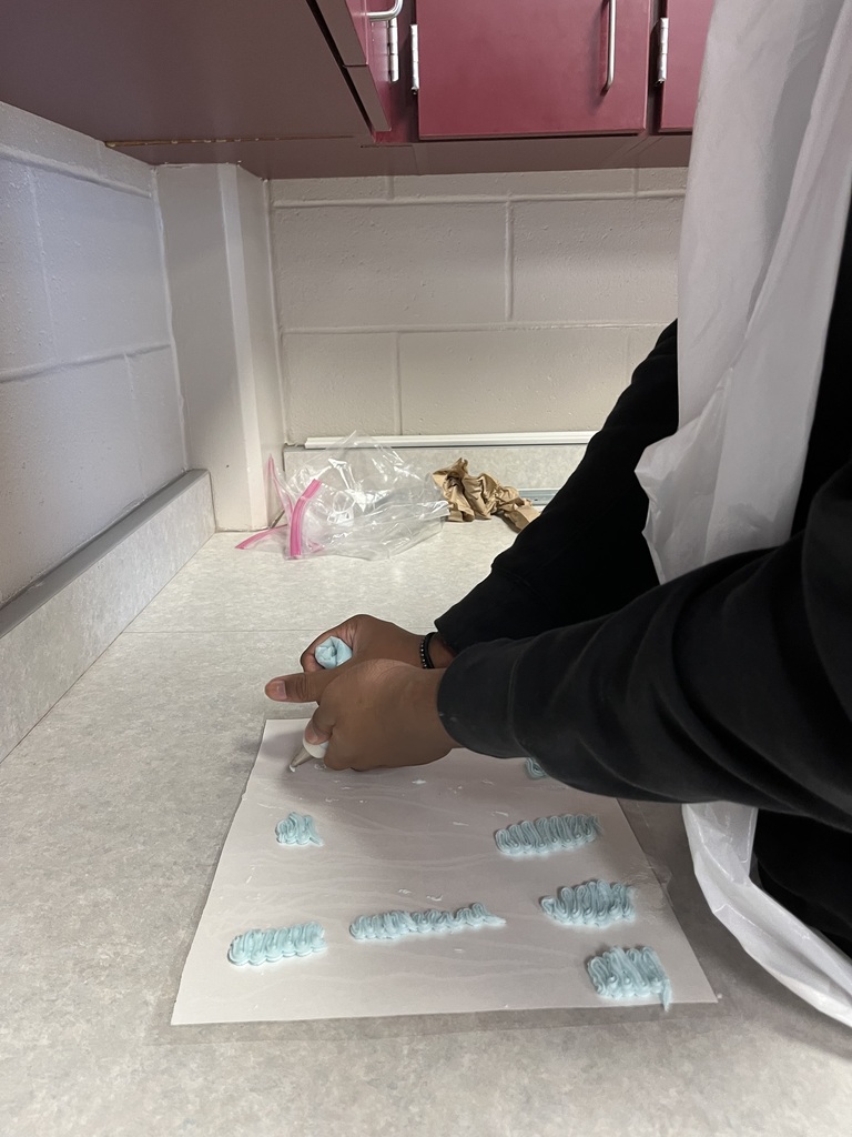 A student in a black shirt and white apron works at a counter, preparing food in a kitchen with brick walls and brown cabinets.