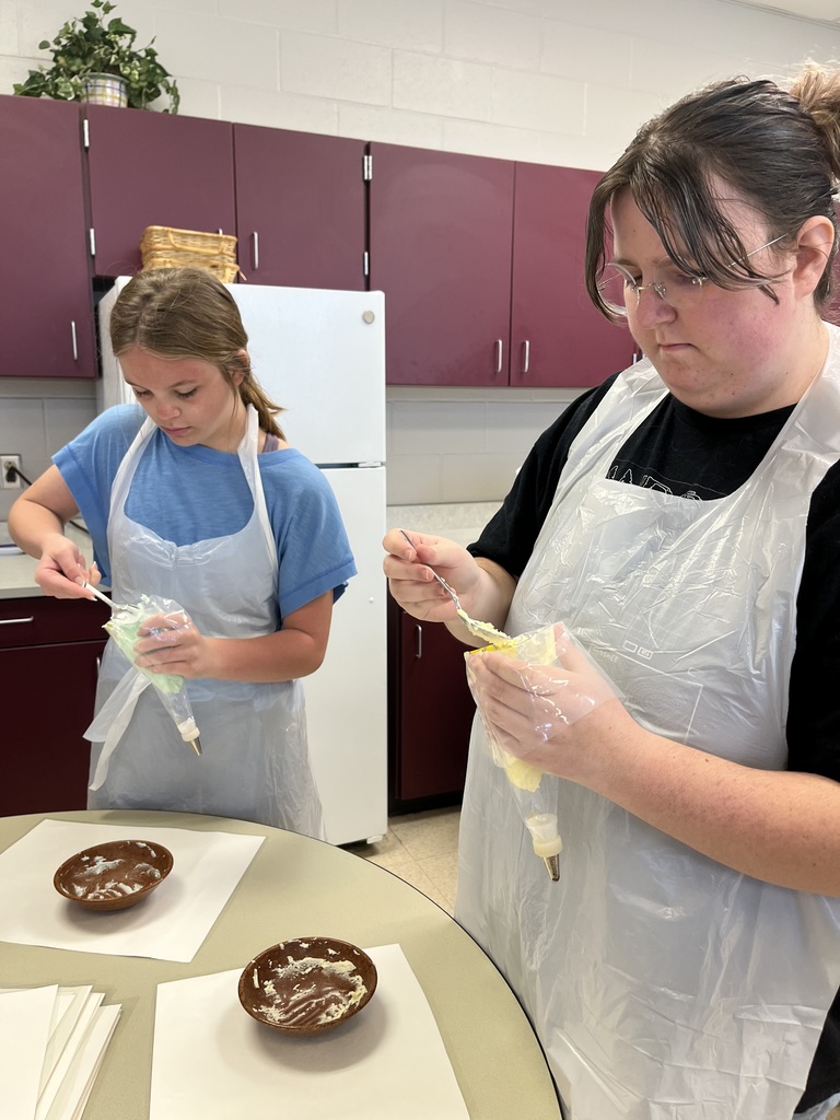 Two students wearing white aprons and glasses focus on preparing food in a kitchen with purple cabinetry