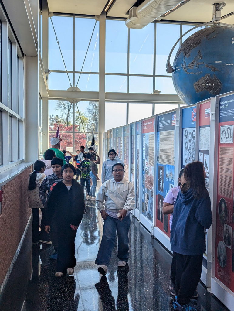 Students walk through a bright museum hallway lined with informational exhibits. Large windows let in natural light, and a large globe display hangs above the exhibit panels.