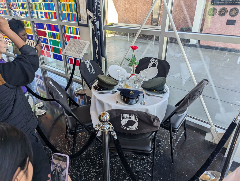 A group of students observes a POW/MIA remembrance table display featuring a white tablecloth, a single red rose, place settings, and military caps. The display is roped off and positioned near a colorful glass wall.