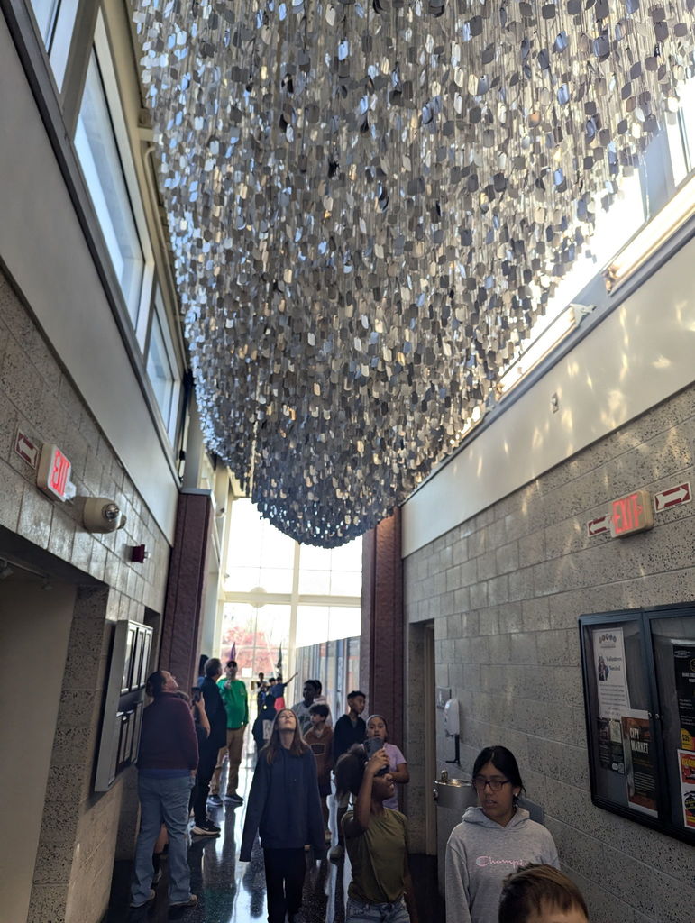 Students and adults walk through a museum corridor while looking up at a large hanging art installation made of reflective metal pieces suspended from the ceiling. Sunlight streams through tall windows at the end of the hallway.