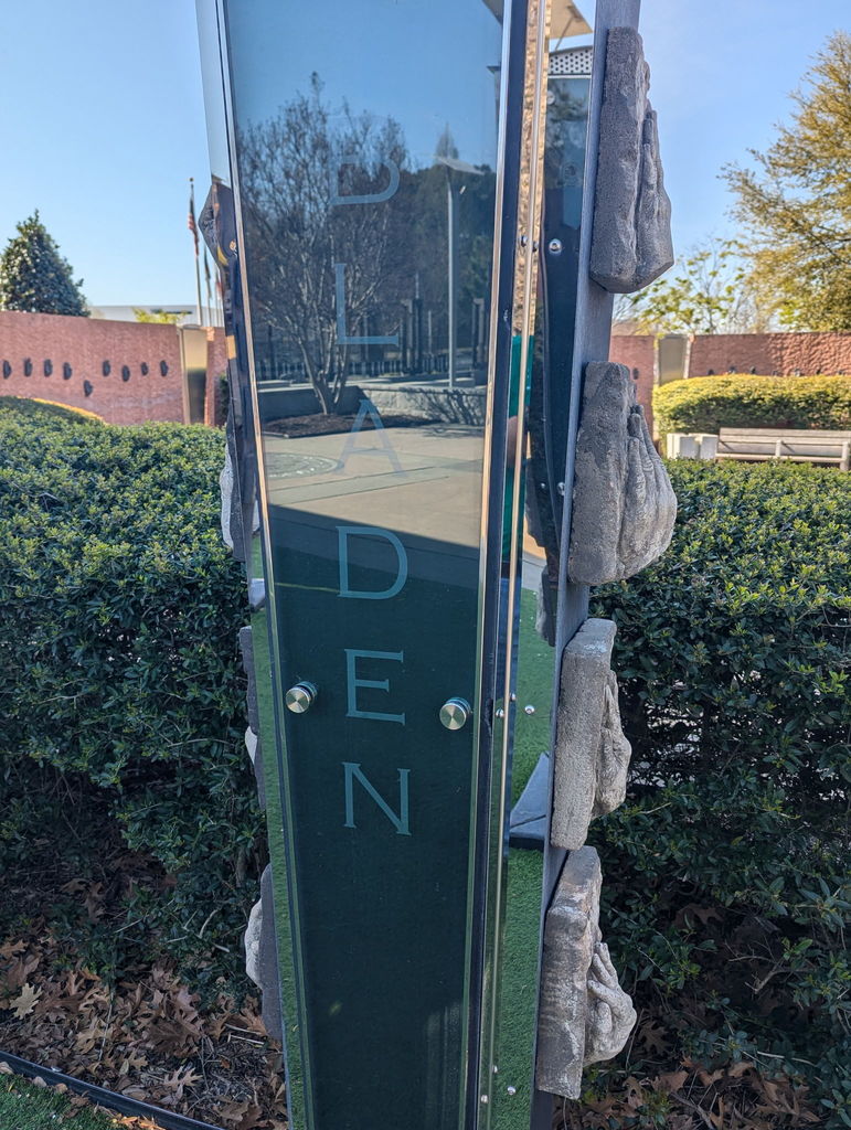 A close-up of a vertical glass and metal monument engraved with the word “BLADEN,” surrounded by greenery and a memorial setting in the background.