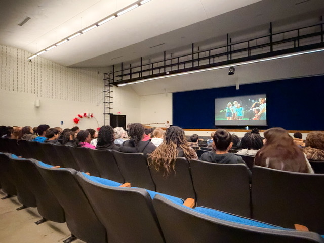 tudents sit in an auditorium watching a presentation on a large screen at the front of the room. Rows of seats are filled as the audience focuses on the stage.
