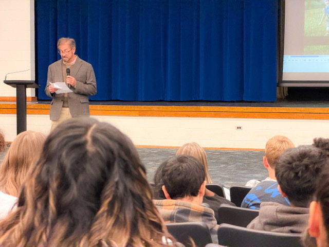 A speaker stands on stage holding a microphone and reading from a paper while students sit in the audience listening attentively.