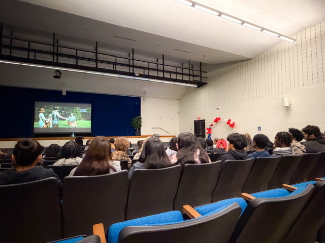 tudents sit in an auditorium watching a presentation on a large screen at the front of the room. Rows of seats are filled as the audience focuses on the stage.