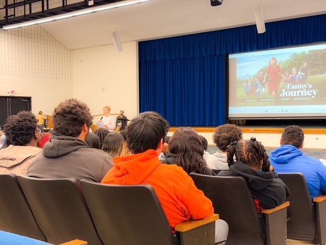 tudents sit in an auditorium watching a presentation on a large screen at the front of the room. Rows of seats are filled as the audience focuses on the stage.