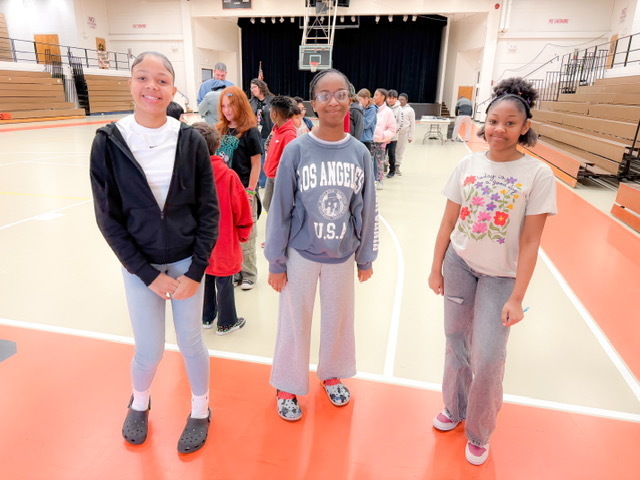 Three students stand in a gym smiling while other students line up behind them for an activity.