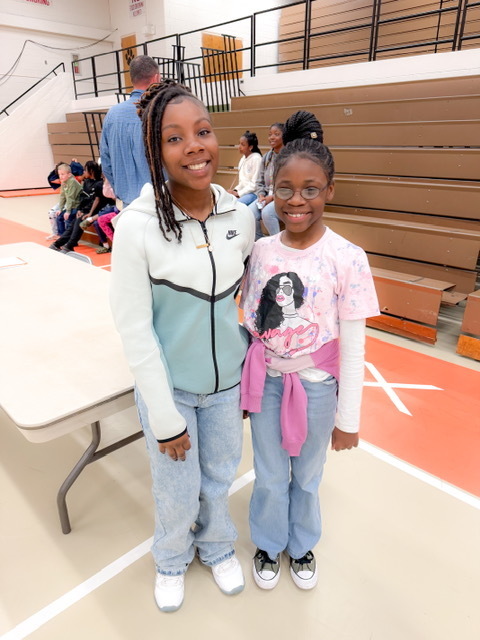 Two students smile and stand side by side in a gym near bleachers, with other students seated in the background.