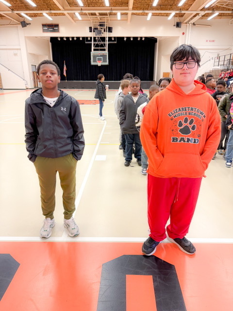 Two students stand at the front of a line in a gym, with more students waiting behind them near the stage.