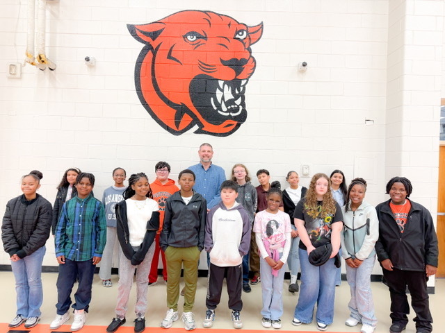 Students and a staff member stand together in a gym for a group photo under a large cougar mascot mural.