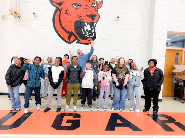 A group of middle school students stand in a gym smiling and posing playfully beneath a large orange cougar mural on the wall.