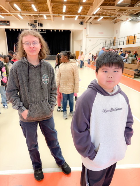 Two students stand at the front of a line in a gym, with more students waiting behind them near the stage.