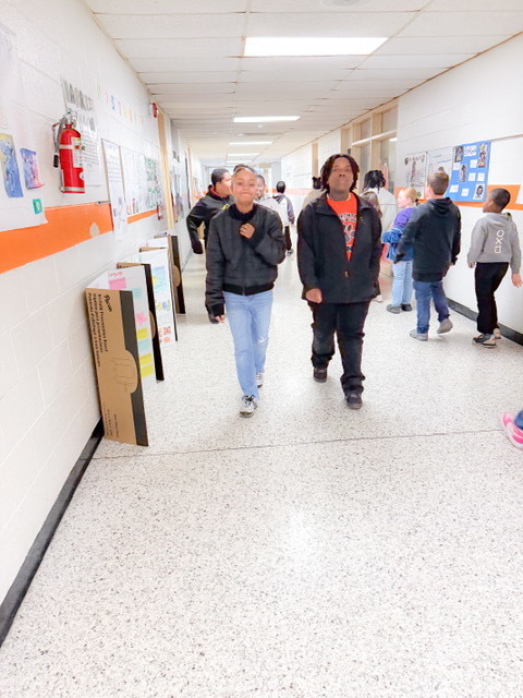 Students walk down a school hallway lined with posters and classroom displays, chatting as they move between classes.
