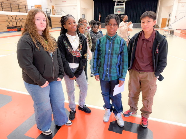 Four students stand together in a gym, posing for a photo while holding papers, with classmates gathered behind them.