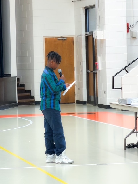 A student stands in a gym holding a microphone and reading from a paper during a presentation.