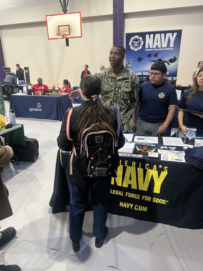 A student with a backpack stands at a U.S. Navy recruitment table featuring brochures and a "Global Force for Good" banner