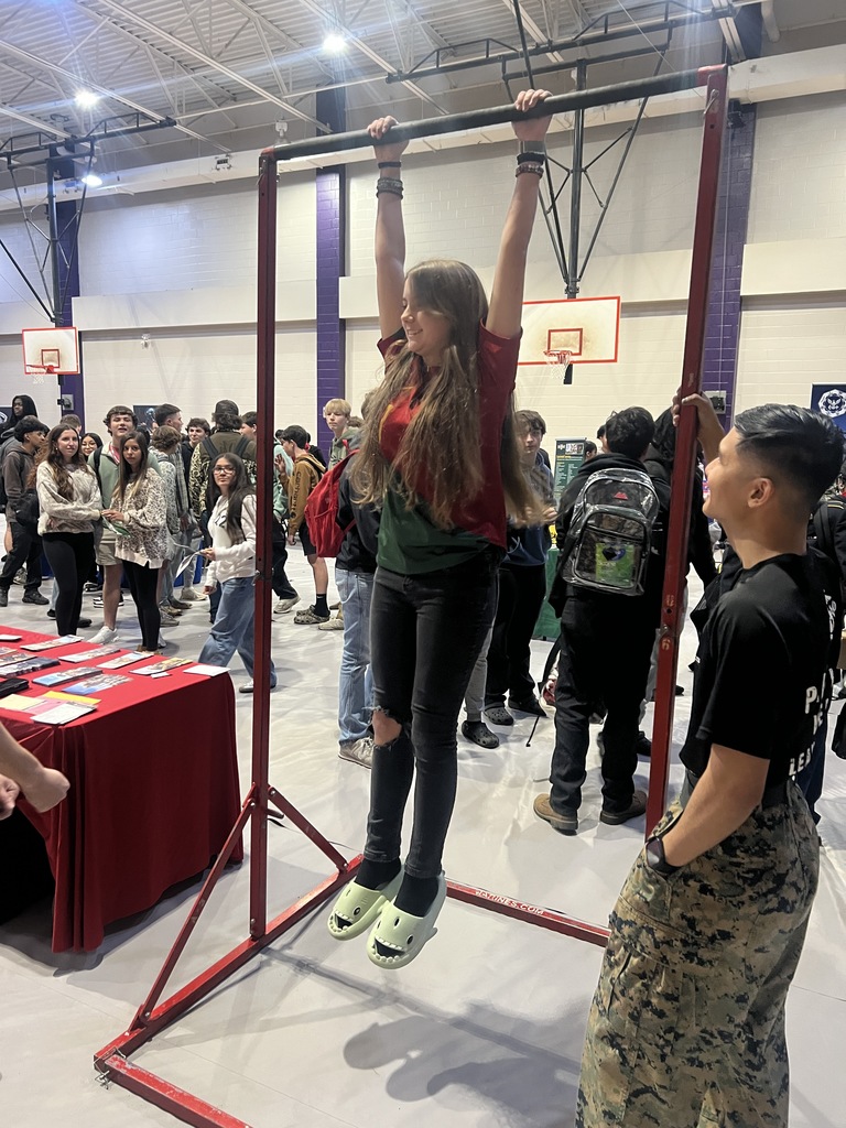 A smiling student hangs from a pull-up bar at a career fair while a crowd of classmates and a recruiter watch in a gym