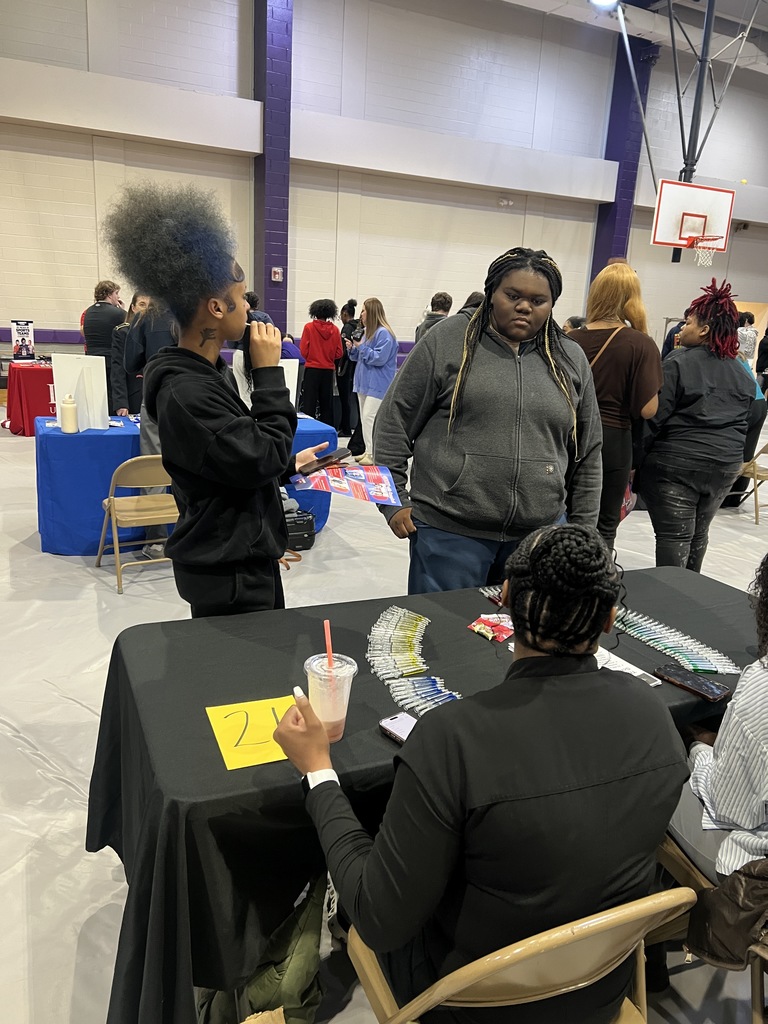 Students gather in a high school gymnasium for a career fair, interacting at various information booths and tables
