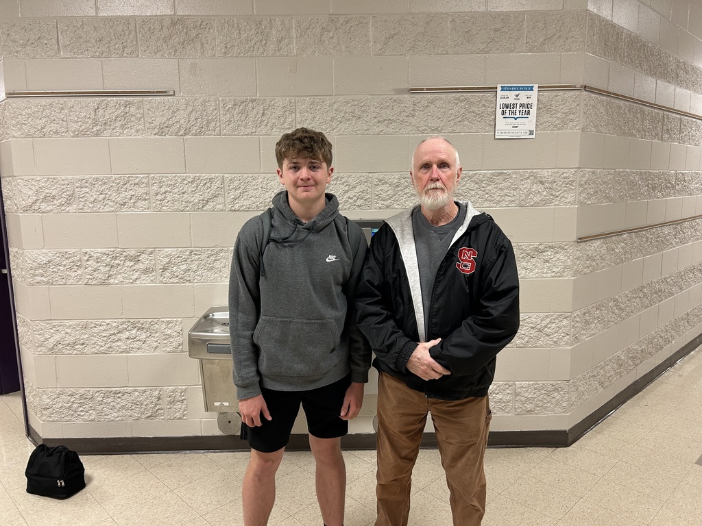A young man in a grey hoodie and an older man in an NC State jacket standing together against a tan brick wall