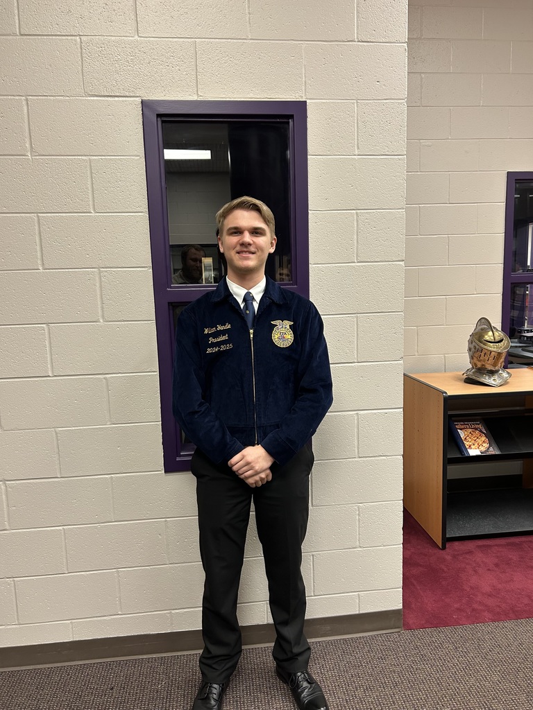 A smiling young man in a blue FFA corduroy jacket standing in front of a window and a tan brick wall.