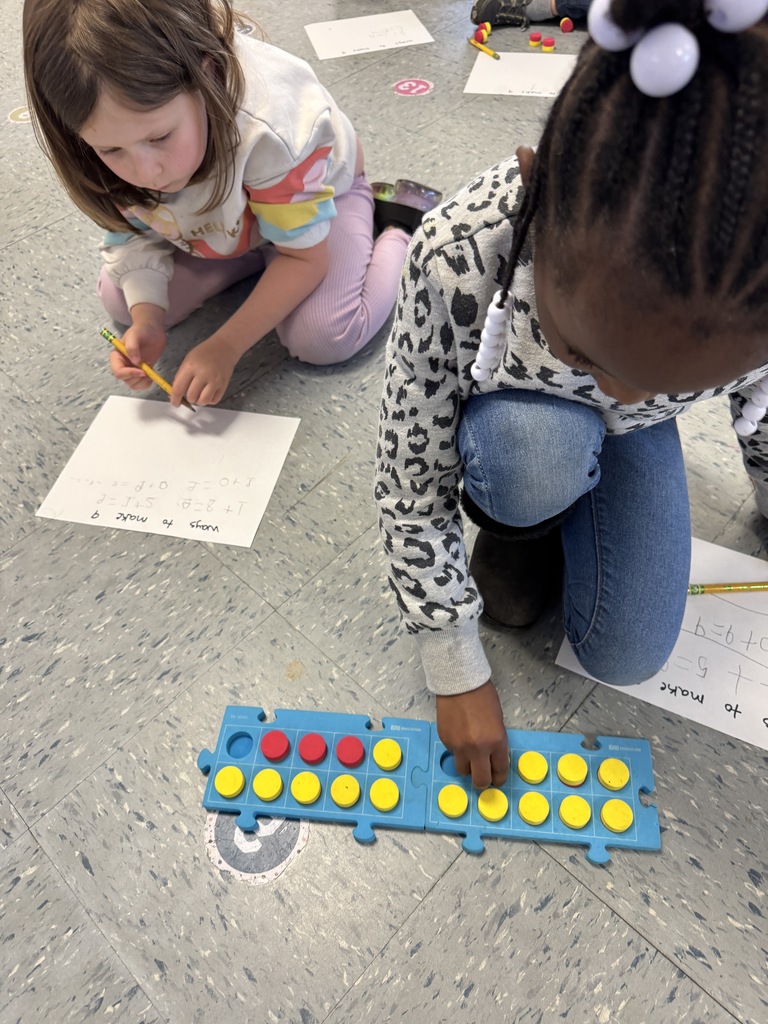 Kindergarten students sit on the floor and at tables using colorful counters and blue math boards to practice number combinations. They write equations on paper as they work together on early addition and number sense skills.