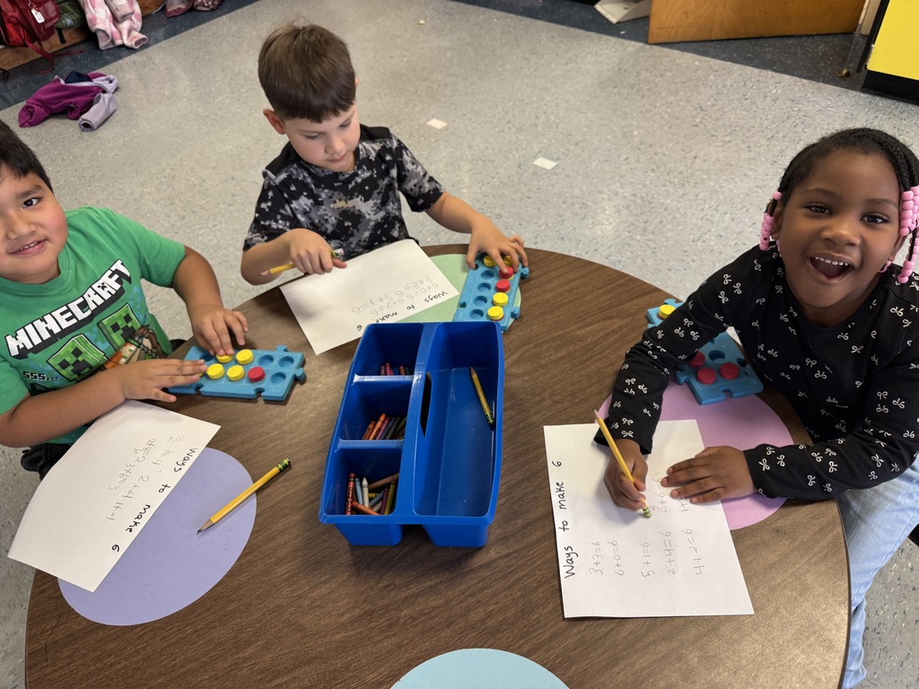 Kindergarten students sit on the floor and at tables using colorful counters and blue math boards to practice number combinations. They write equations on paper as they work together on early addition and number sense skills.