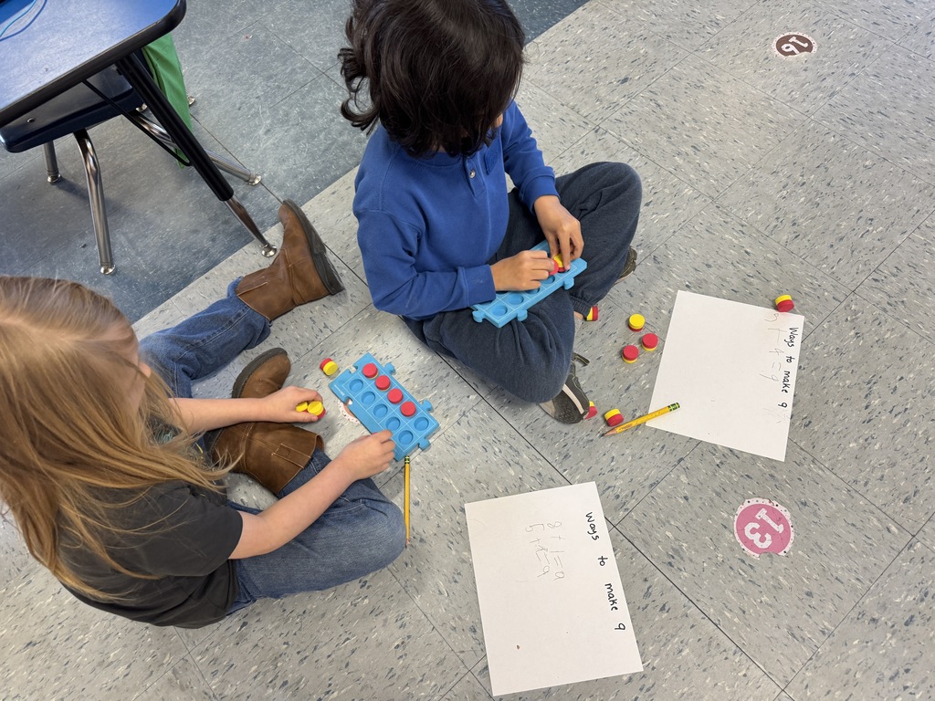 Kindergarten students sit on the floor and at tables using colorful counters and blue math boards to practice number combinations. They write equations on paper as they work together on early addition and number sense skills.