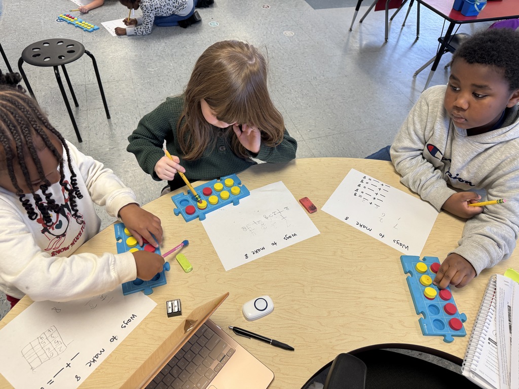 Kindergarten students sit on the floor and at tables using colorful counters and blue math boards to practice number combinations. They write equations on paper as they work together on early addition and number sense skills.