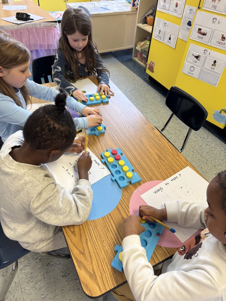 Kindergarten students sit on the floor and at tables using colorful counters and blue math boards to practice number combinations. They write equations on paper as they work together on early addition and number sense skills.