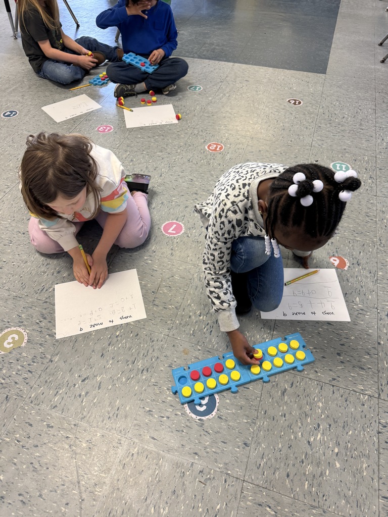 Kindergarten students sit on the floor and at tables using colorful counters and blue math boards to practice number combinations. They write equations on paper as they work together on early addition and number sense skills.