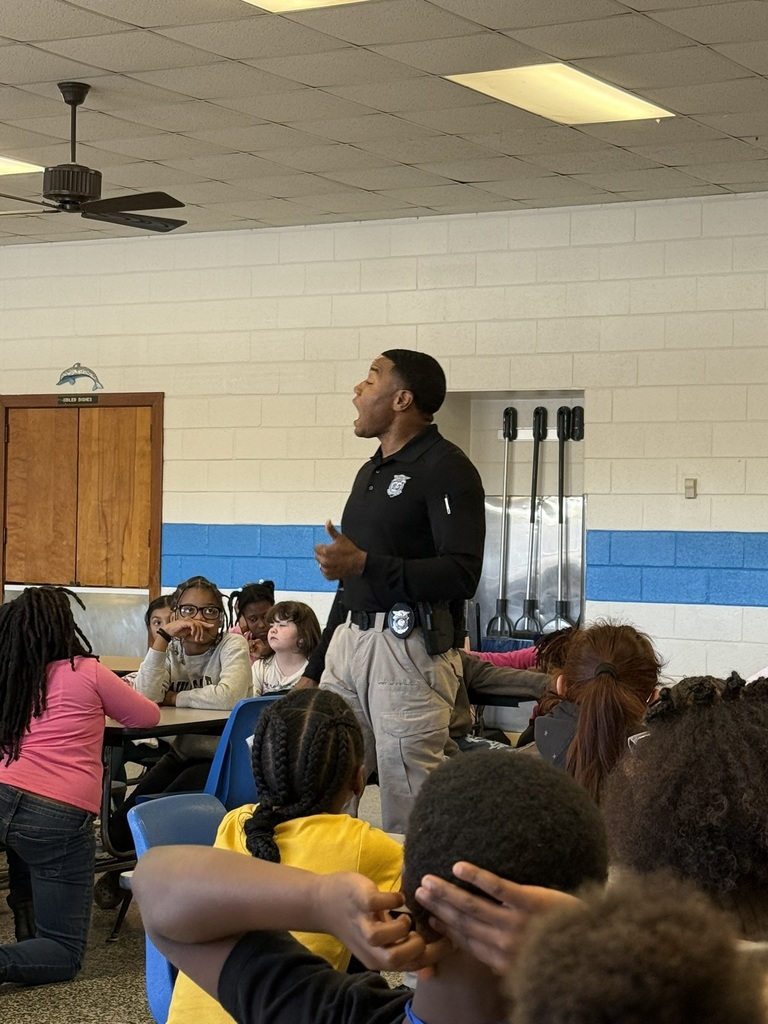 A North Carolina Highway Patrol officer stands and speaks to a group of fourth grade students seated in a cafeteria. Students listen attentively as he shares a message about responsibility, perseverance, and making positive choices.