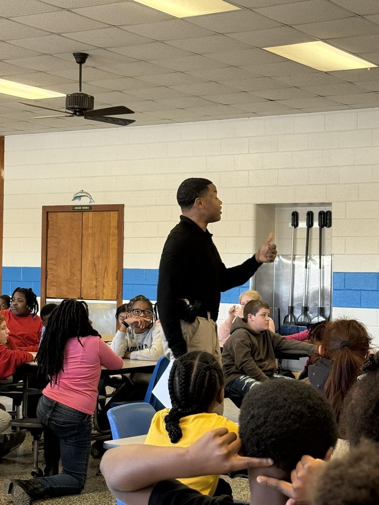 A North Carolina Highway Patrol officer stands and speaks to a group of fourth grade students seated in a cafeteria. Students listen attentively as he shares a message about responsibility, perseverance, and making positive choices.