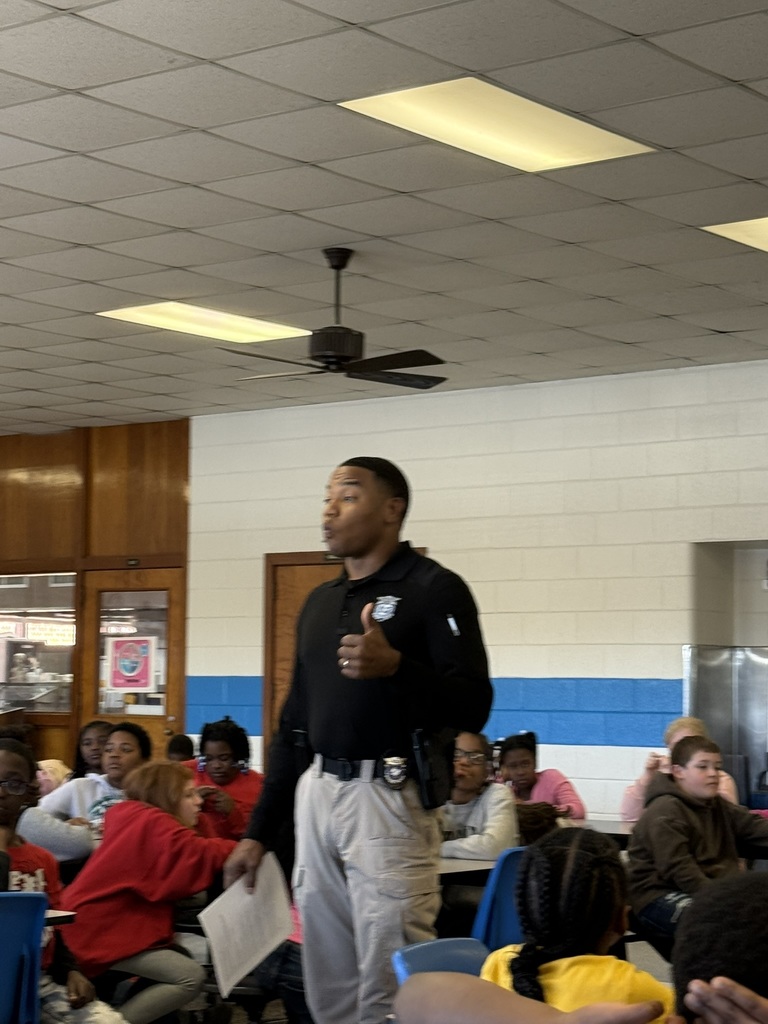 A North Carolina Highway Patrol officer stands and speaks to a group of fourth grade students seated in a cafeteria. Students listen attentively as he shares a message about responsibility, perseverance, and making positive choices.
