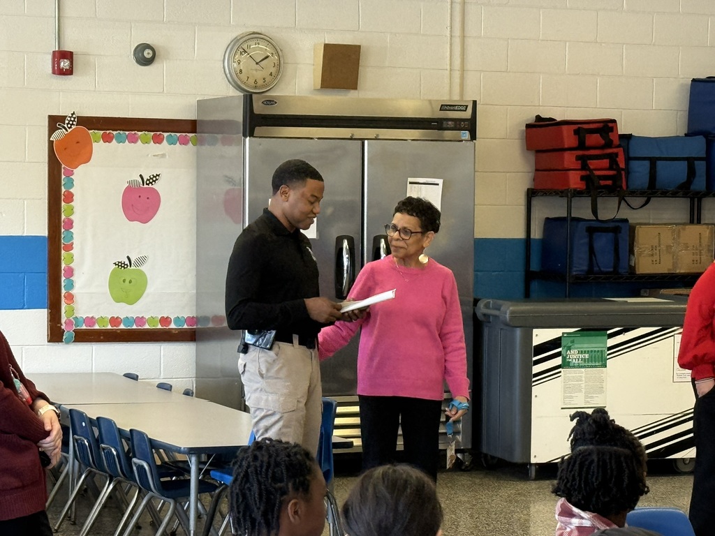 A North Carolina Highway Patrol officer stands and speaks to a group of fourth grade students seated in a cafeteria. Students listen attentively as he shares a message about responsibility, perseverance, and making positive choices.
