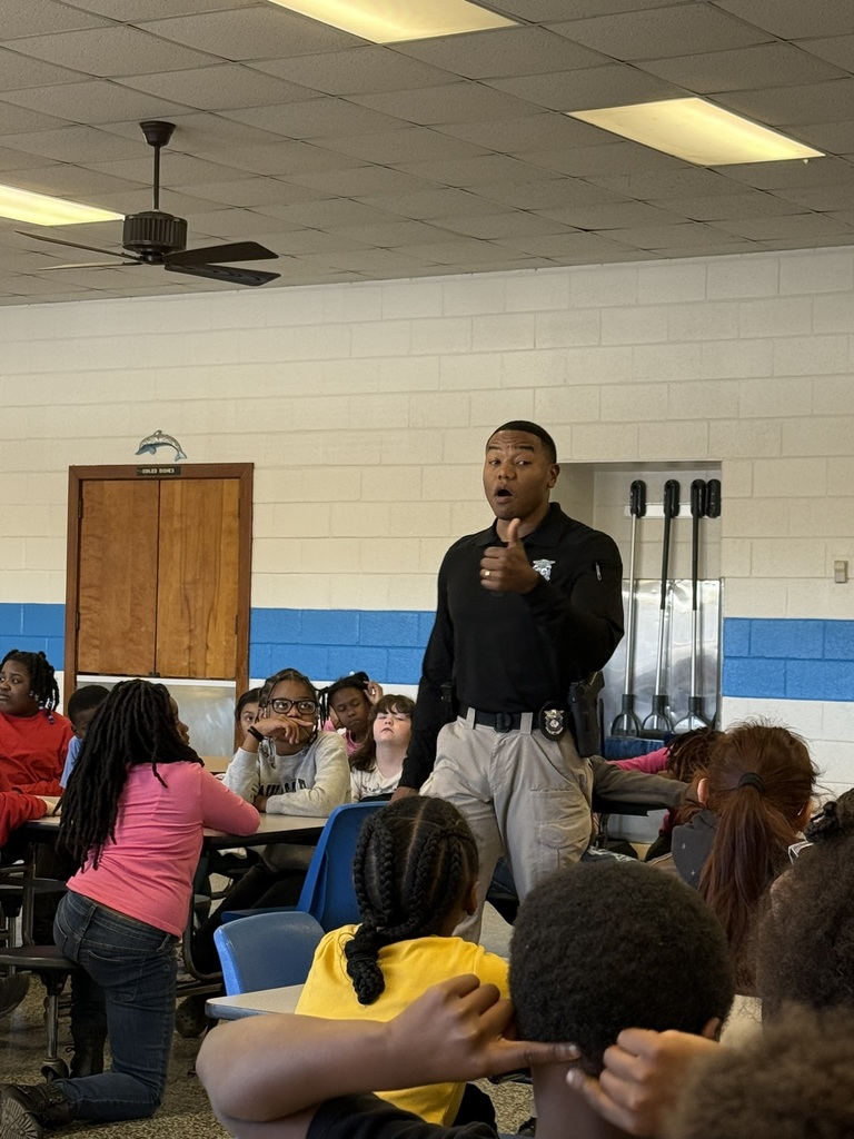 A North Carolina Highway Patrol officer stands and speaks to a group of fourth grade students seated in a cafeteria. Students listen attentively as he shares a message about responsibility, perseverance, and making positive choices.
