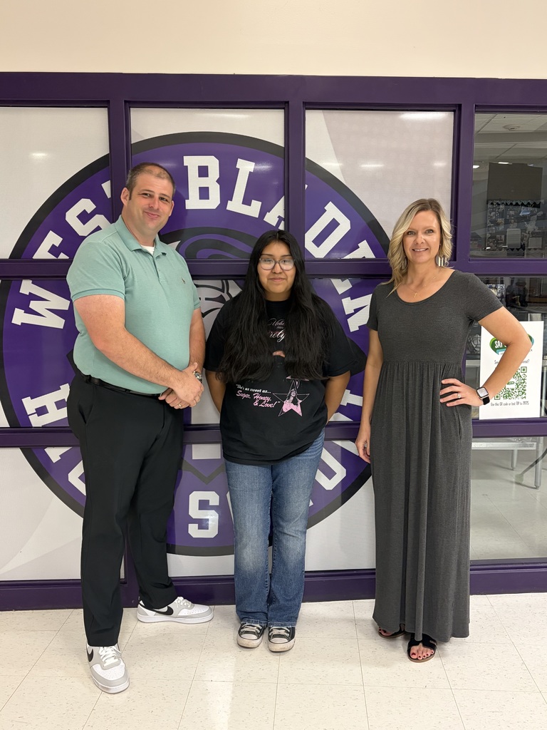 A man in a green shirt, a girl in a black t-shirt, and a woman in a grey dress stand smiling in front of a purple and white "West Bladen" wall graphic