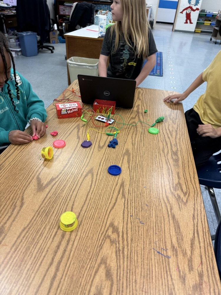 Young students work together at a table using Play-Doh and Makey Makey to build circuits and experiment with sound on a laptop.