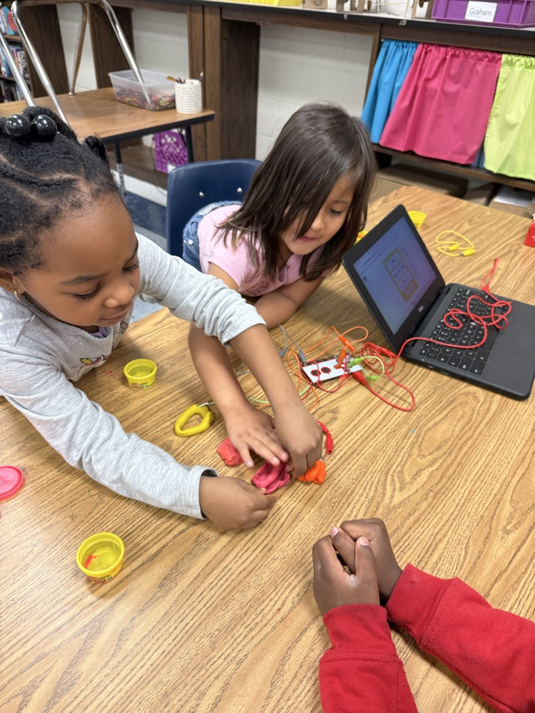 Young students work together at a table using Play-Doh and Makey Makey to build circuits and experiment with sound on a laptop.