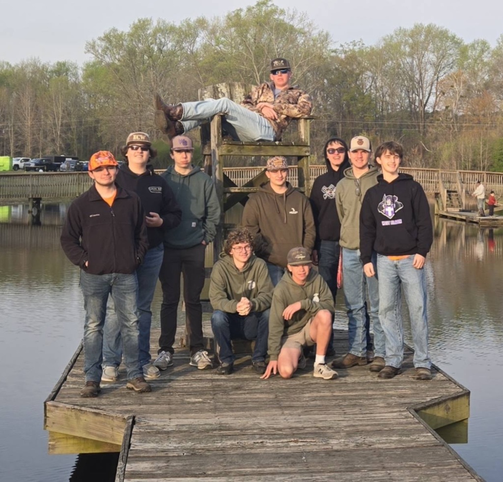 A group of nine male students and one instructor posing for a photo outdoors near a wooden boardwalk and pond. One student is sitting playfully in a high wooden chair in the center, while the others stand in two rows in front of him wearing hoodies and hats. The background features a line of budding spring trees and a clear sky