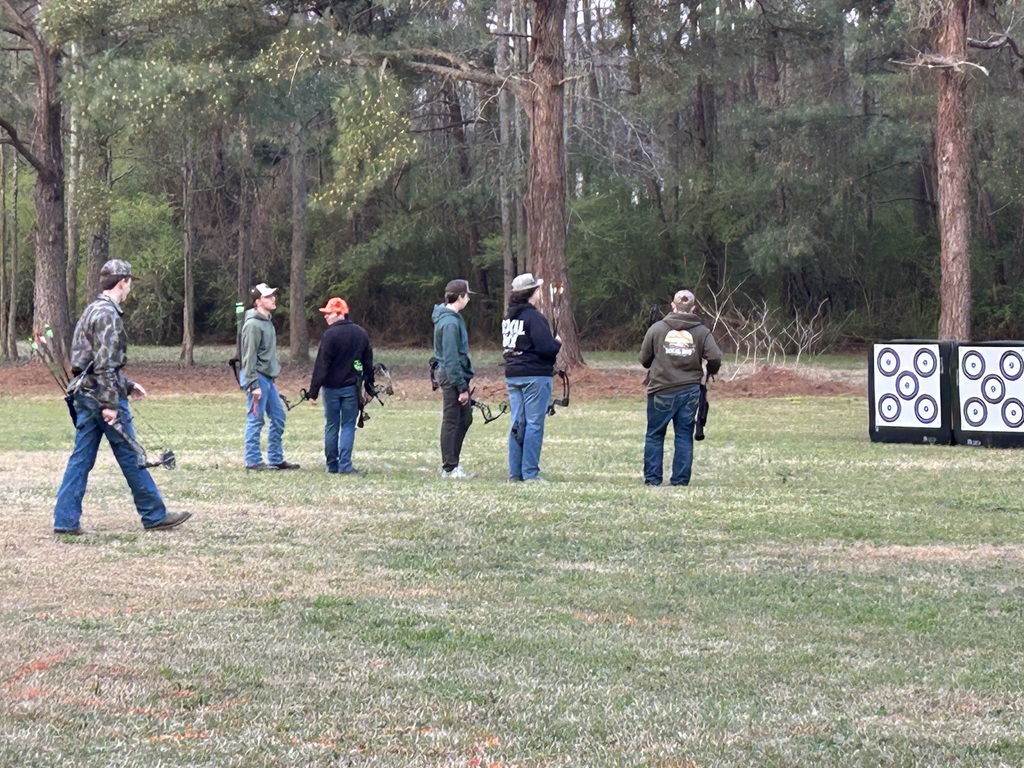A group of student athletes and instructors participating in an outdoor archery session on a grassy field, with several archers holding bows and standing near target blocks backed by a pine forest
