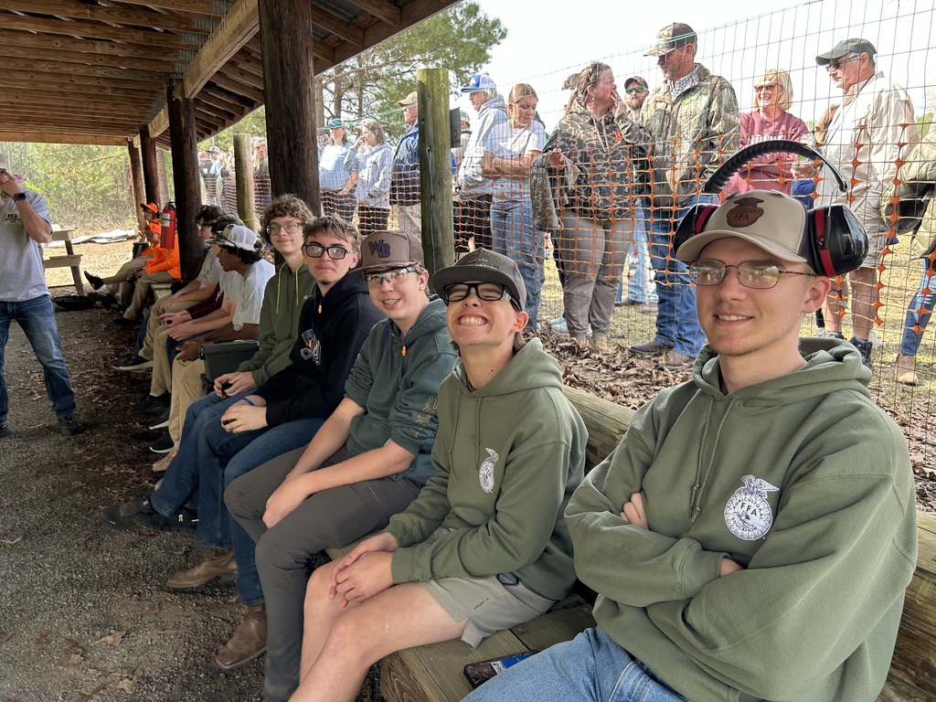 A group of five smiling high school students, wearing casual outdoor attire and safety ear protection, sit together on a wooden bench at an outdoor shooting range. In the background, other students and instructors are standing near a safety fence under a wooden pavilion
