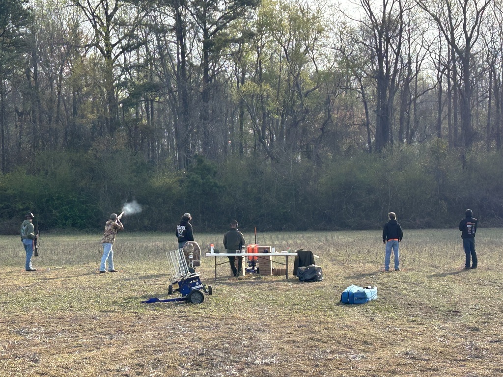 A group of people practicing target shooting with shotguns in an open grassy field bordered by a dense line of bare winter trees. One individual in a camouflage jacket is captured mid-shot with a small puff of white smoke coming from the barrel, while others stand nearby observing or waiting their turn