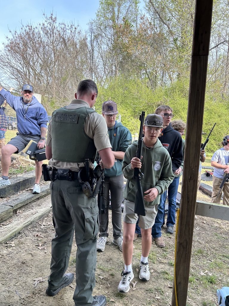 A group of high school students and instructors wearing camouflage and outdoor gear stand together on a grassy field during a target shooting or wildlife education event. One student in the foreground is seen from the back, wearing a "NCWRC" (North Carolina Wildlife Resources Commission) vest, while others in the background are holding equipment near a wooded treeline under a clear sky